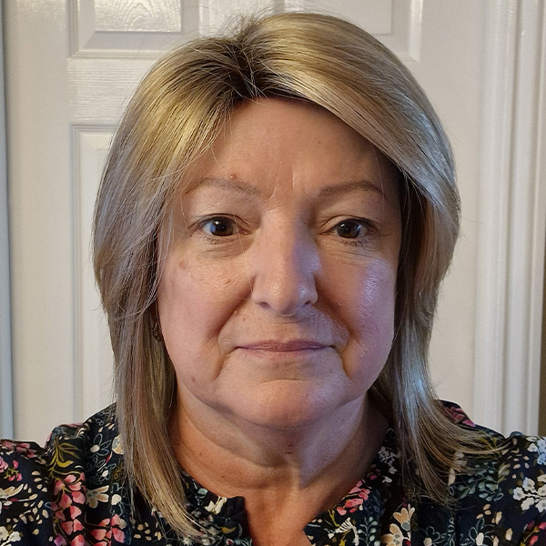 Woman with a shoulder-length, straight honey-blonde bob with a side parting, wearing a dark floral top