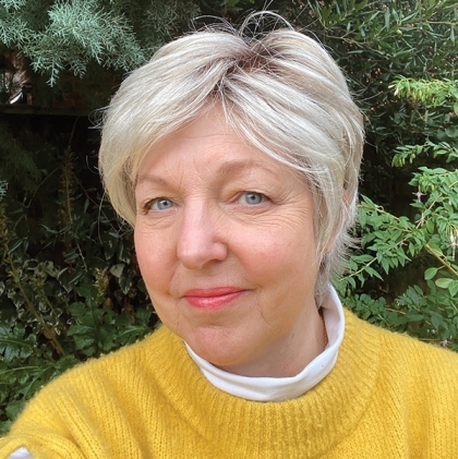 Woman with a short, layered platinum blonde pixie cut and side-swept fringe, wearing a yellow jumper in a garden