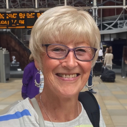 Smiling older woman with a short, feathered light grey style wig and purple-framed glasses