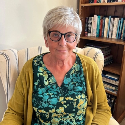 Woman with a short, soft grey crop and dark-rimmed glasses, wearing a green cardigan, seated by a bookcase
